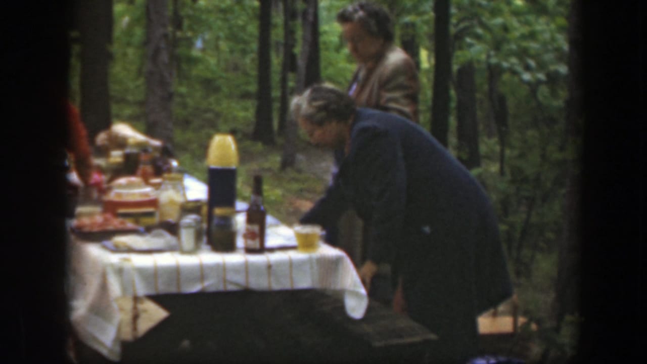 Adults gather by a picnic table with food and bottles in a wooded area.
