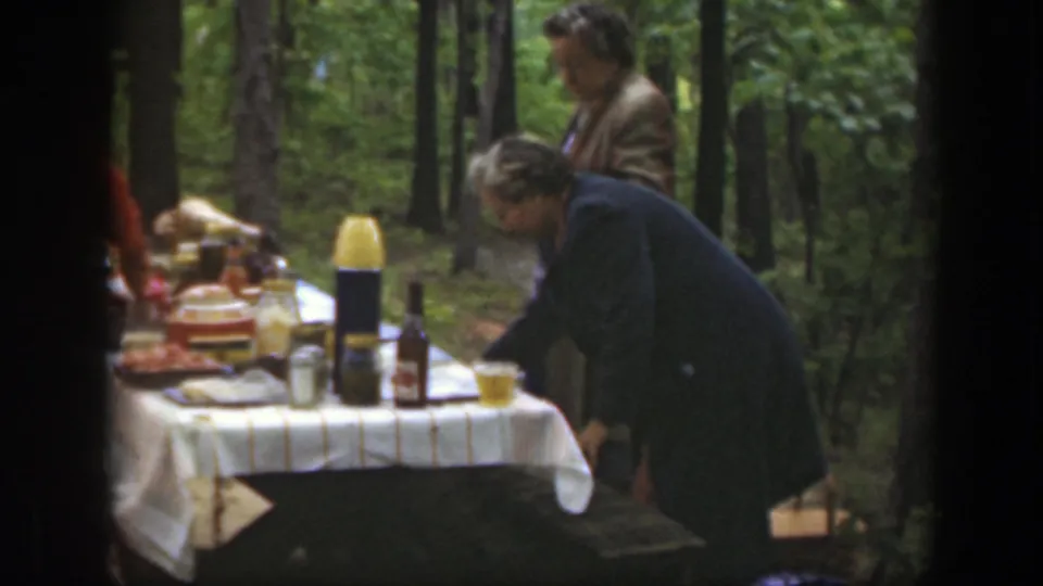 1949 Massachusetts home-movie frame showing adults around a cloth-covered picnic table in the woods.