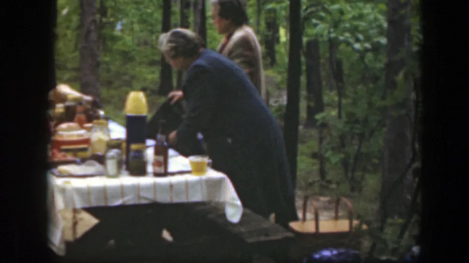 1949 Massachusetts picnic scene with a woman bending over a table of food and drinks in a wooded setting.