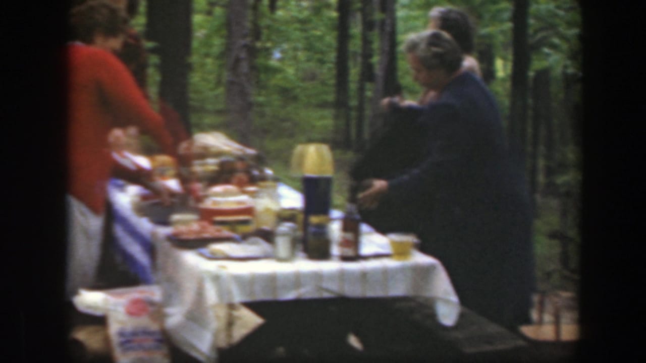 1949 Massachusetts home-movie still of adults preparing or serving food at a wooded picnic table.