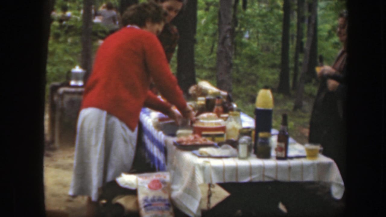 1949 Massachusetts picnic frame with a woman in a red sweater serving from a table amid trees.