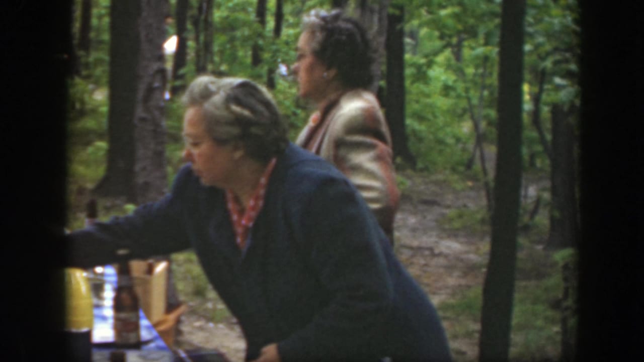 Two women near a picnic table in a wooded park, one reaching toward bottles at the table edge.