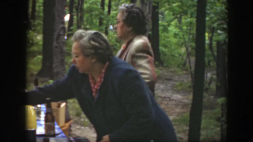 1949 Massachusetts home-movie frame of women beside a picnic table in a wooded park, with bottles and dishes partly visible.
