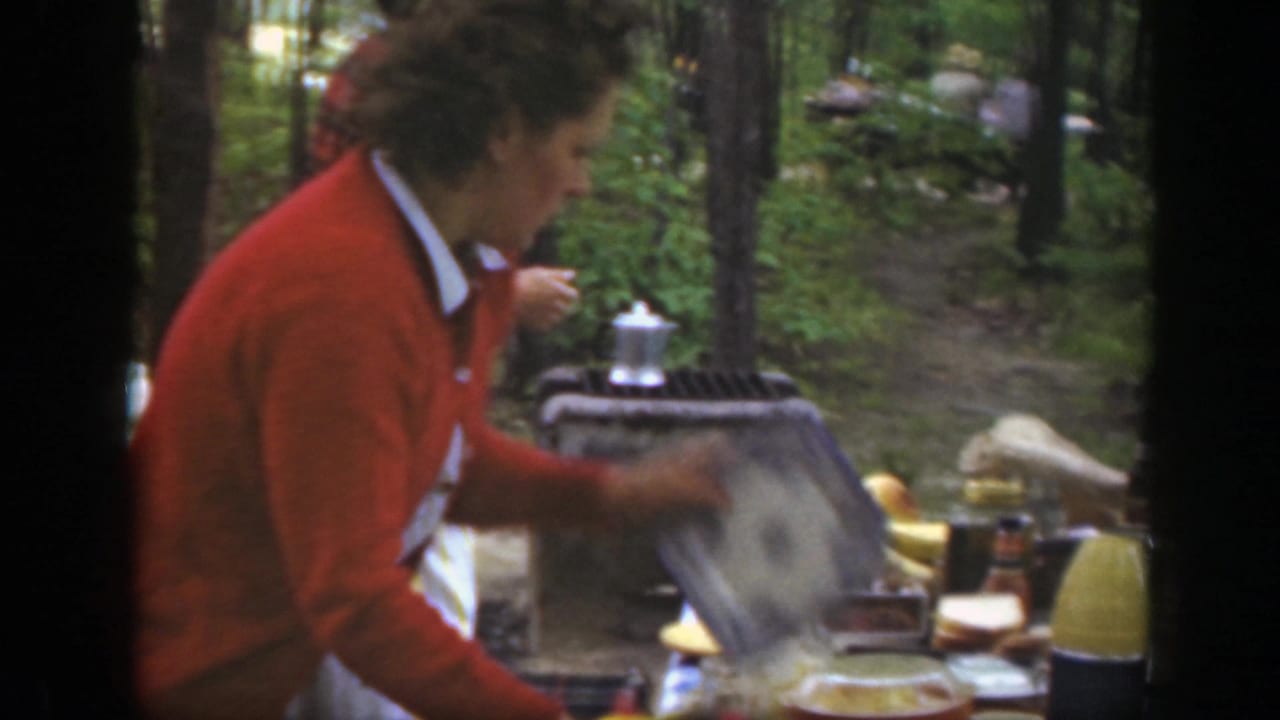 1949 Massachusetts frame showing a woman in a red cardigan working at a picnic table with dishes and a yellow thermos nearby.