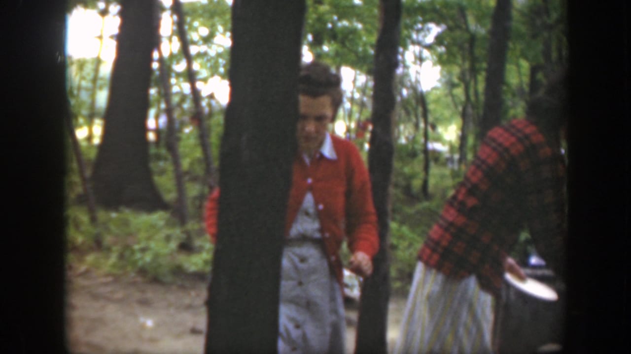 1949 Massachusetts home-movie frame with two women in a wooded campsite, one in a red cardigan and one near cookware.