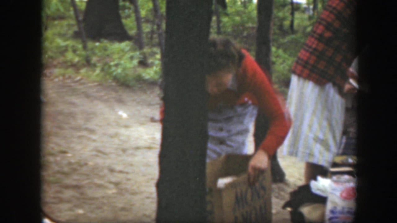 1949 Massachusetts frame with a woman handling a cardboard box beside plates and supplies in a wooded campsite.