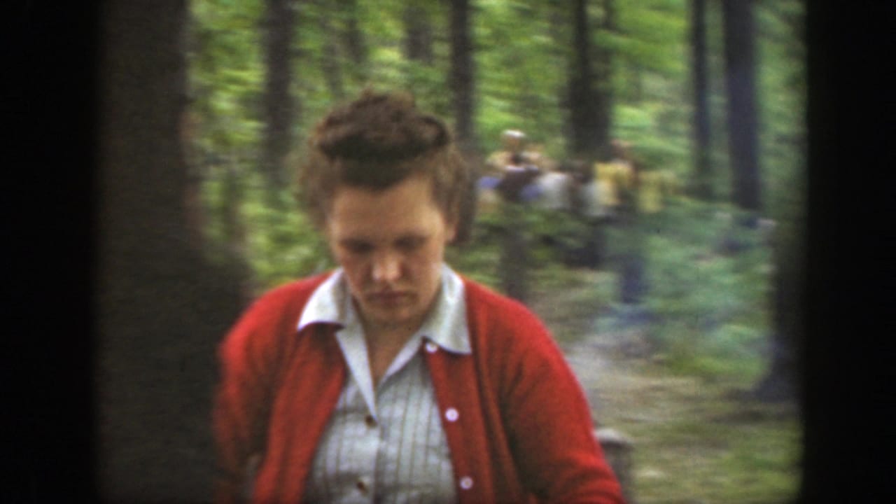 Woman in a red cardigan stands near trees with people gathered in the background.