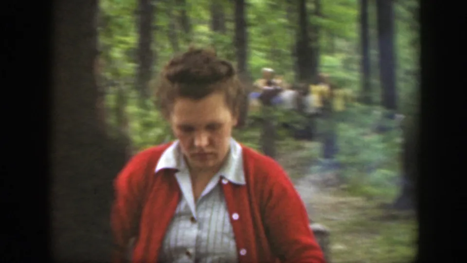 In metadata-supported 1949 Massachusetts footage, a woman in a red cardigan appears in a wooded clearing with adults behind her.