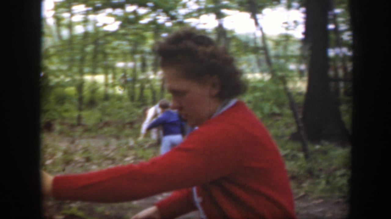 The woman in a red cardigan reaches downward in a Massachusetts wooded setting while other people stand farther back.