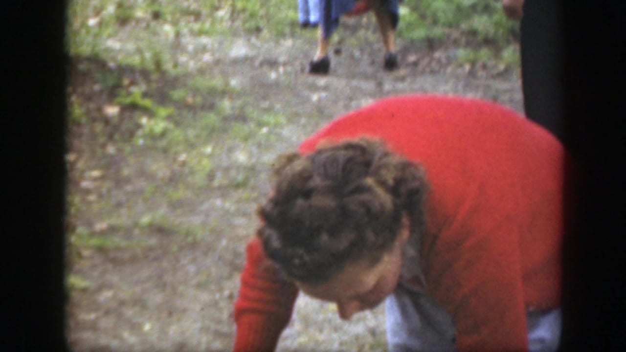A woman in a red cardigan bends toward the ground on a wooded path, with other adults partly visible in the background.