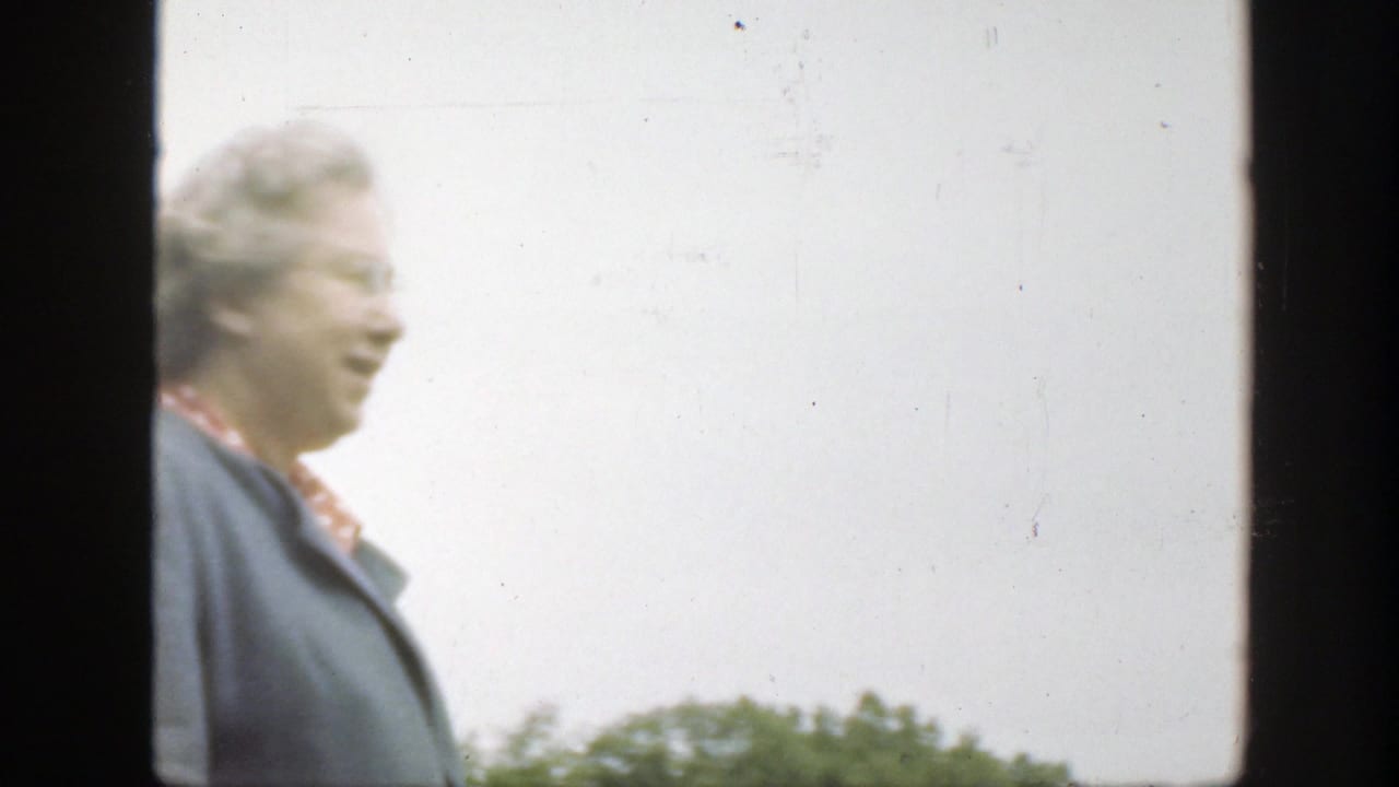 Older woman in a blue jacket smiles outdoors beside a bright sky and trees.