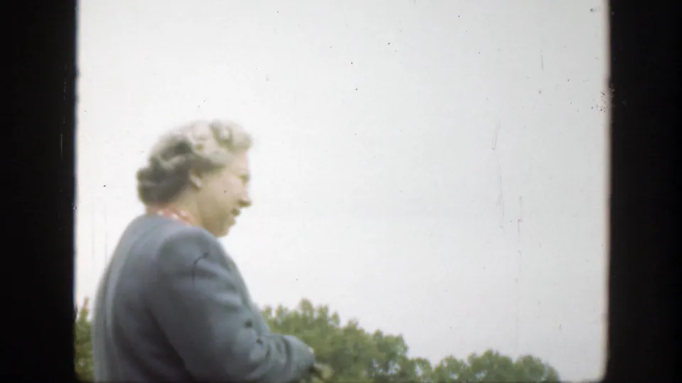 The older woman, wearing a blue jacket and red scarf, looks down or to the side in a soft 8mm outdoor frame.