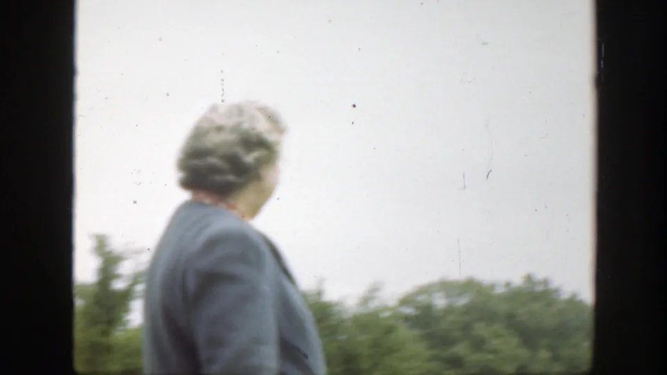 The woman turns away from camera in the outdoor Massachusetts home movie, with green trees and pale sky behind her.