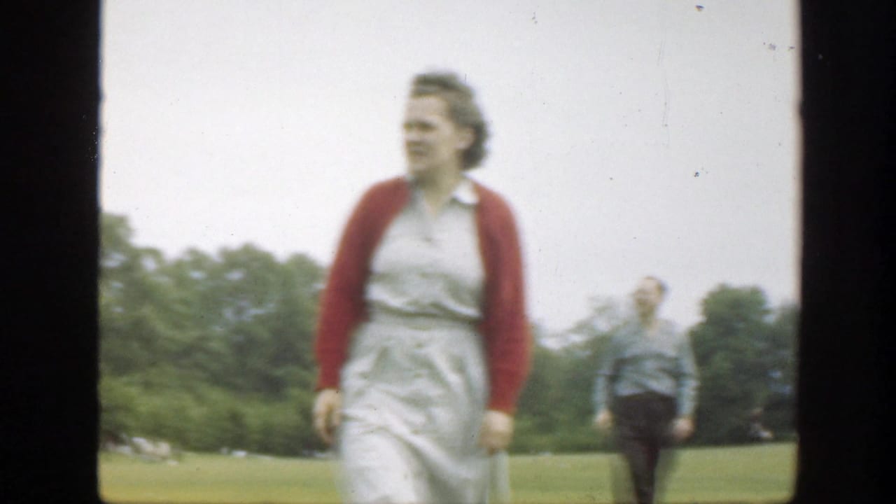 Woman in a white dress and red cardigan walking in a grassy field with a man behind her.