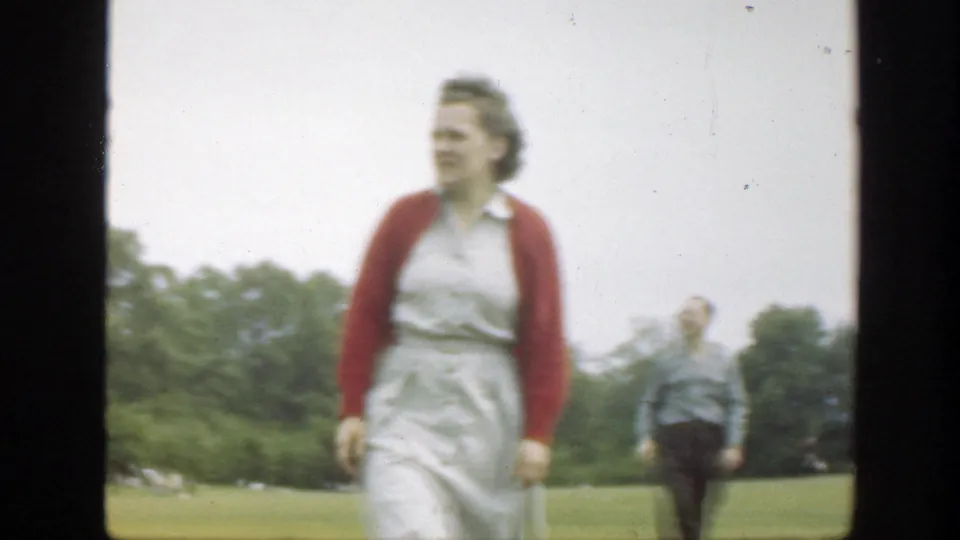In 1949 Massachusetts home-movie footage, a woman in a white dress and red cardigan walks across a grassy field with a man in the distance.