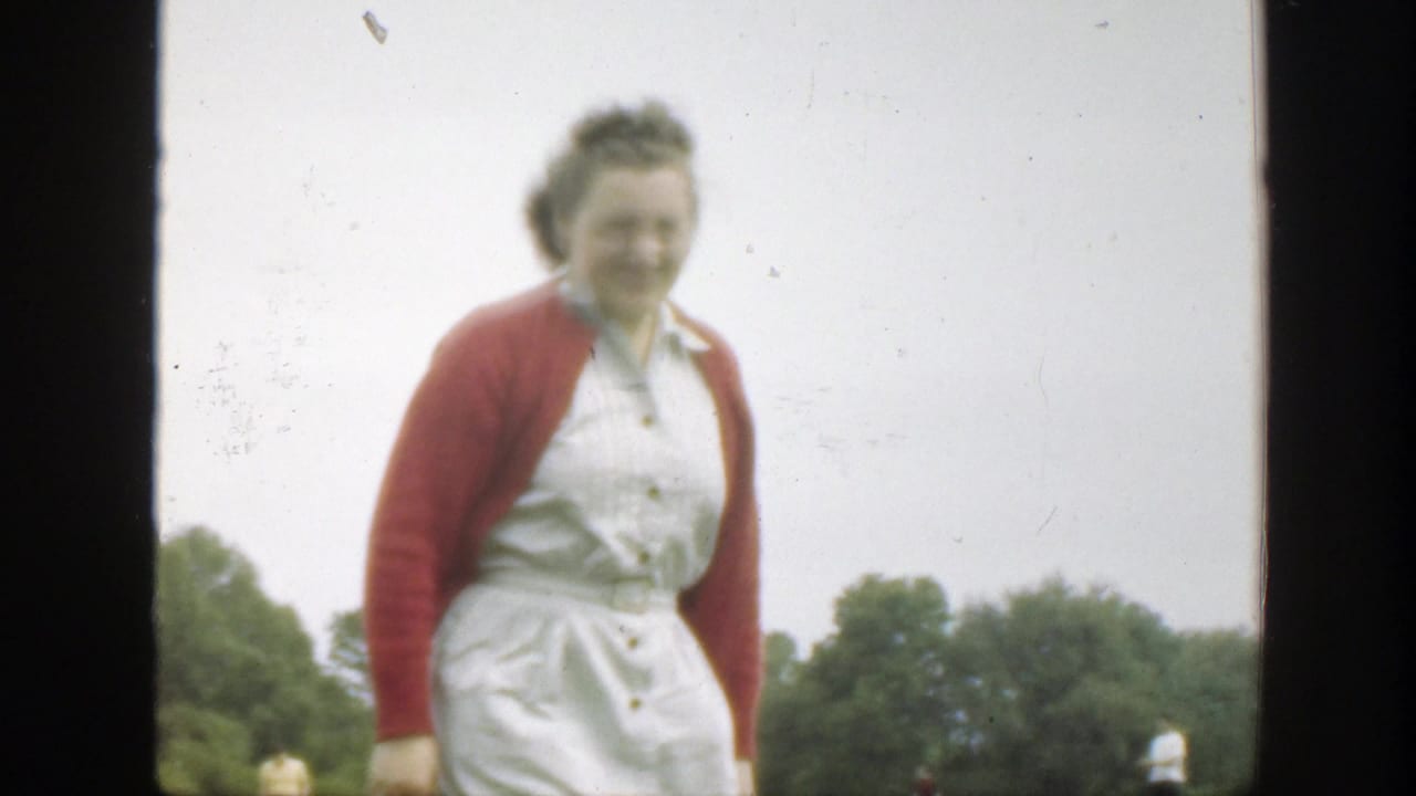 The woman in the red cardigan and white dress smiles while walking nearer to the camera in a Massachusetts field.