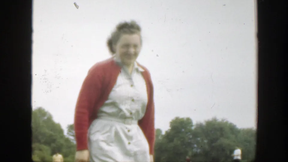The woman in the red cardigan and white dress smiles while walking nearer to the camera in a Massachusetts field.