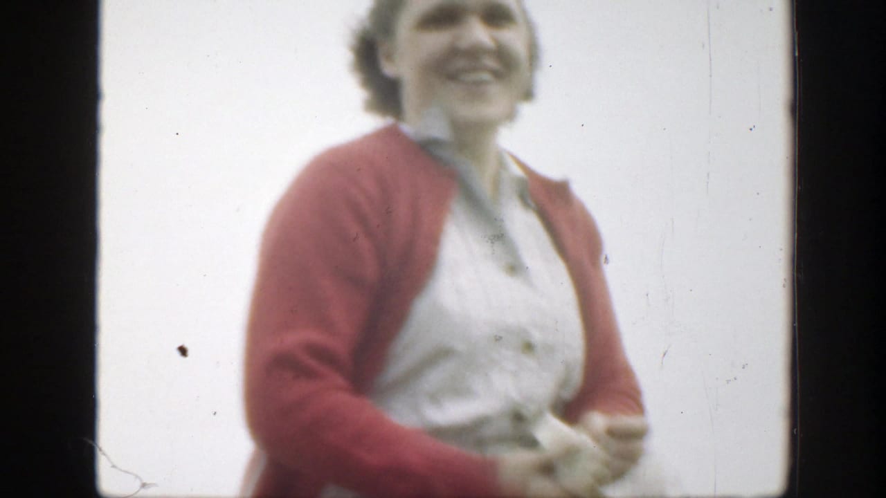 A close, soft-focus view shows the smiling woman in a red cardigan and white dress as she passes near the 8mm camera.