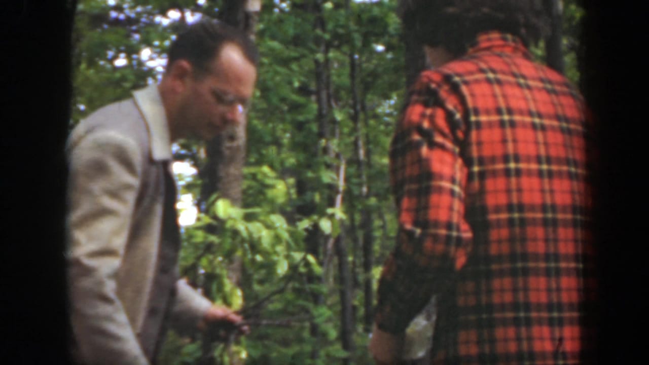 1949 Massachusetts 8mm frame of two adult men in woods, with a man in a light jacket bending beside kindling and another in a red plaid shirt.