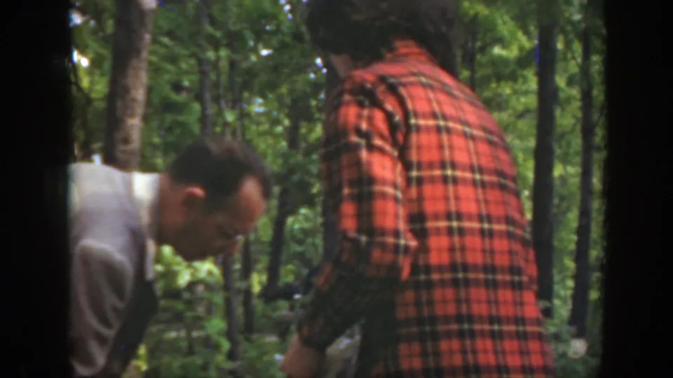 In a Massachusetts wooded setting, a man in a light jacket bends toward the ground while a red-plaid-shirted man stands nearby.