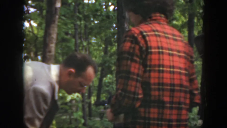 1949 Massachusetts home-movie frame showing two adult men close to the forest floor during outdoor fire or kindling preparation.