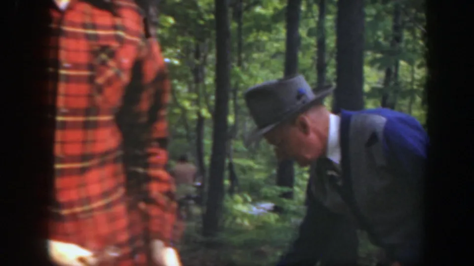 A third adult man in a fedora appears in the Massachusetts woods as the red-plaid-shirted man remains nearby during fire preparation.