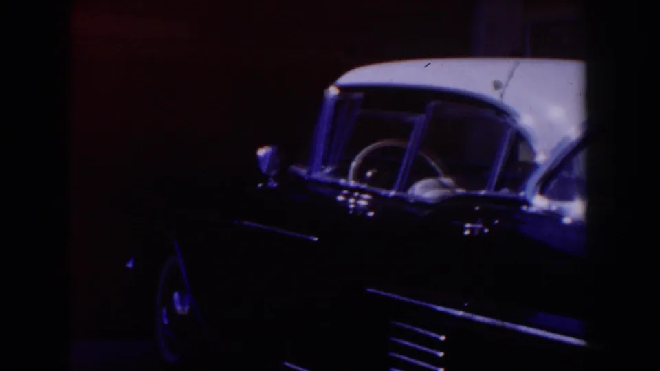 1962 Glendale home-movie frame showing a dark classic car with a white roof, windshield, chrome trim, and steering wheel in dim red light.