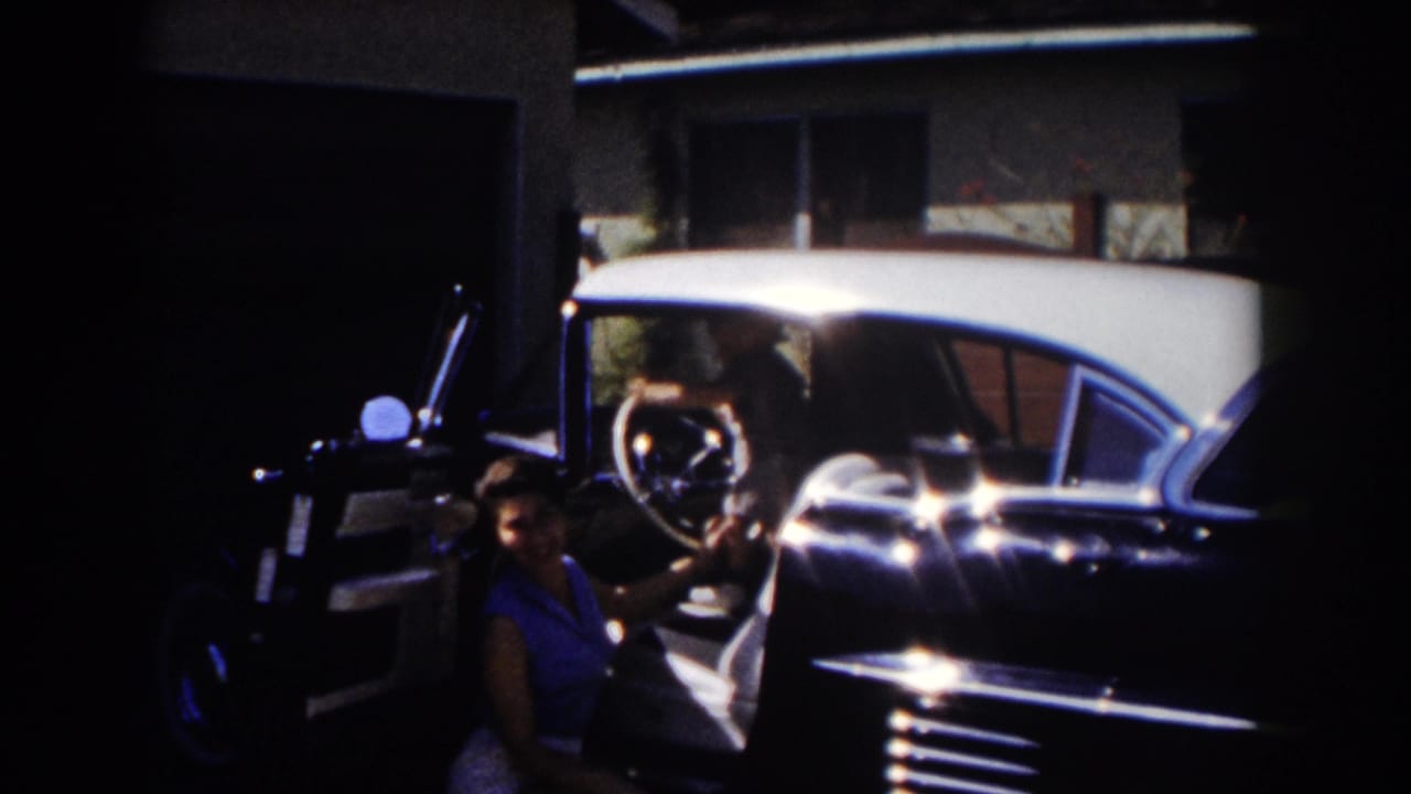 Woman in a blue sleeveless top stands beside an open car door in a shaded driveway.