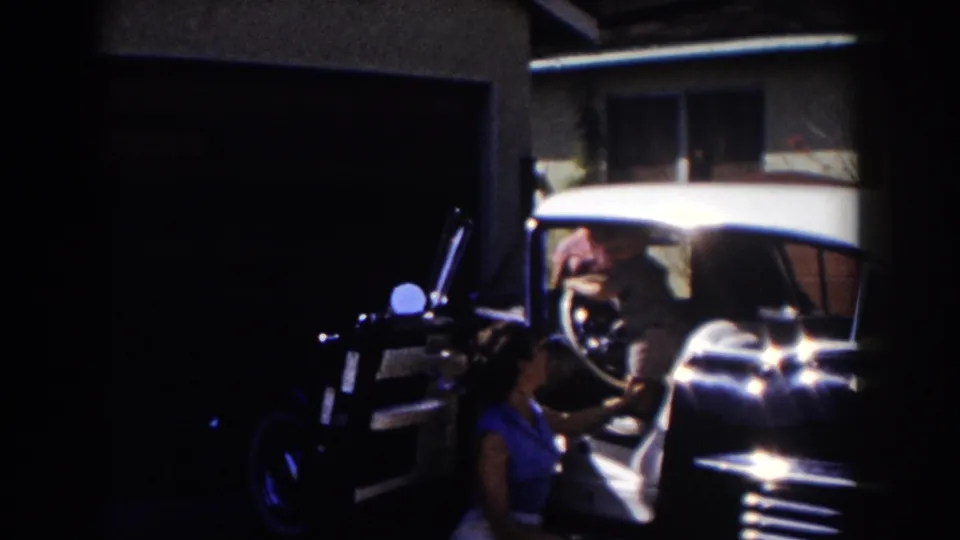A woman in blue stands near the open door of a mid-century car as someone sits at the steering wheel in a Glendale driveway.