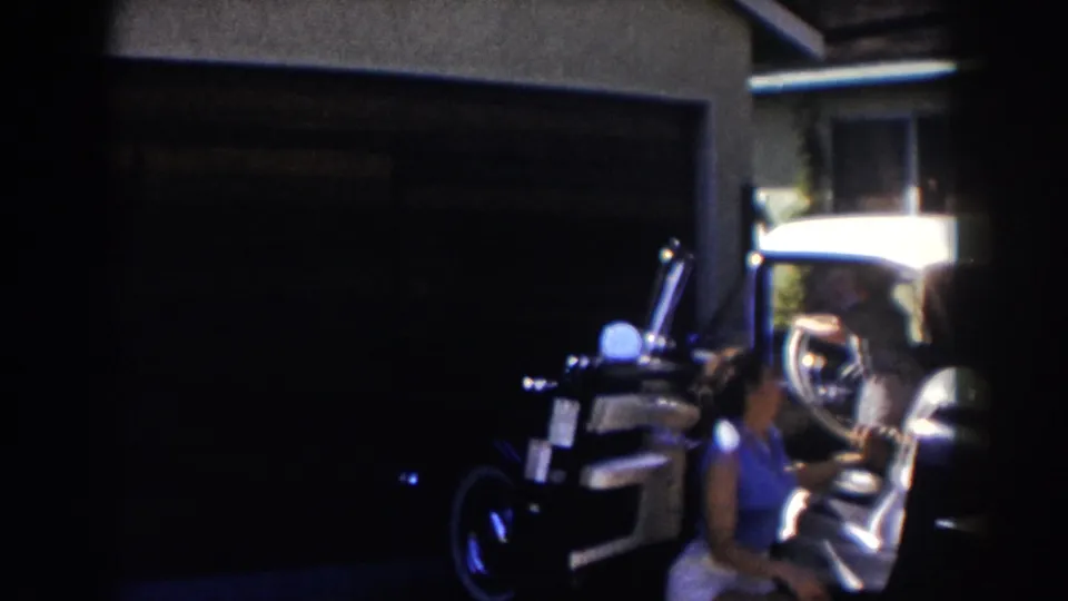 The woman is lower beside the open car door, with the steering wheel and bright two-tone car body visible in the driveway.