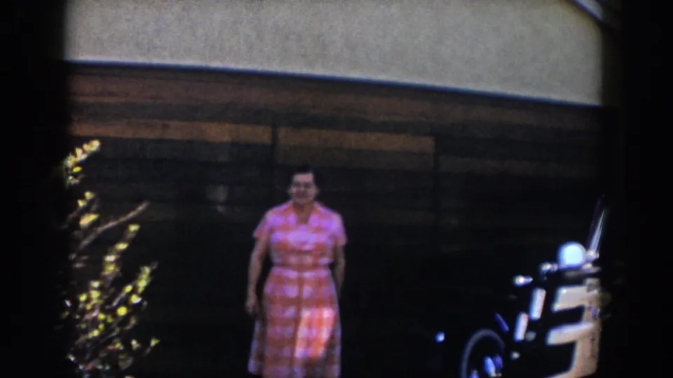 An adult in a pink patterned dress stands in sunlight outside the garage while the white classic car remains partly visible.