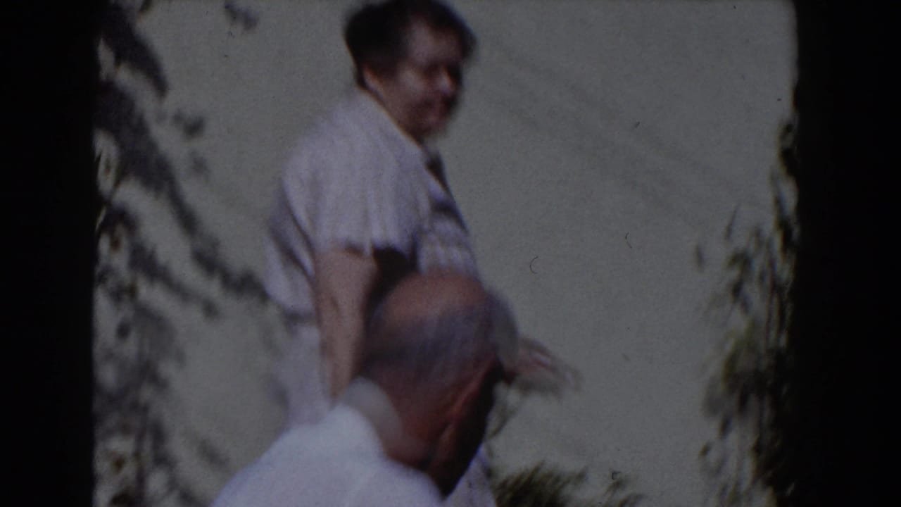 Older woman and older man stand near a backyard wall and plants.
