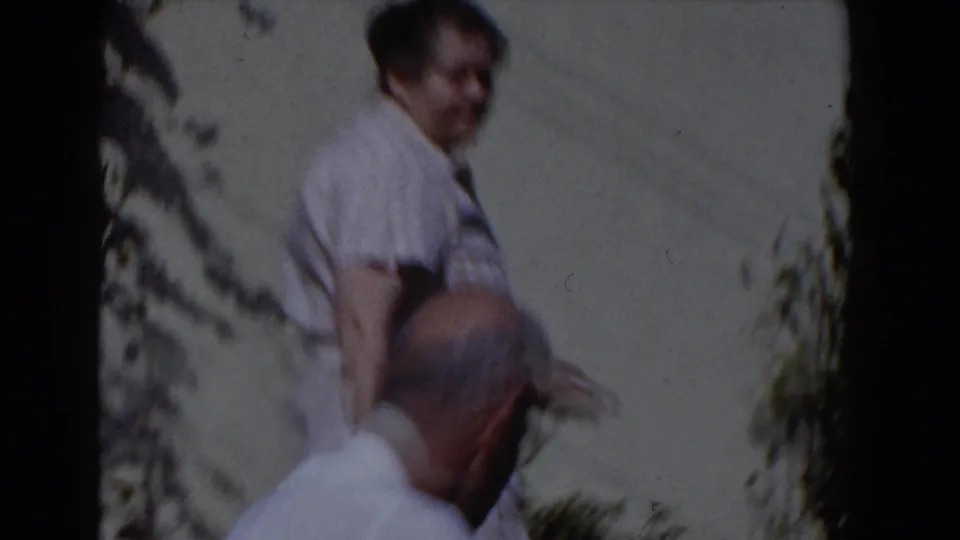 The older couple appears in the Glendale backyard, with the woman standing near the wall as the man turns in the foreground.