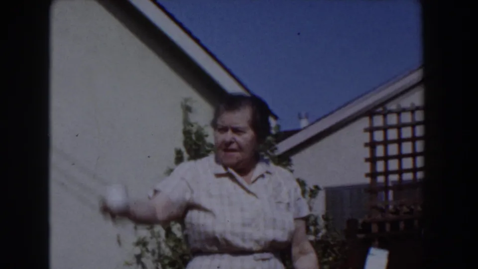 An older woman in a light checkered dress moves her arm to toss a white ball in the 1962 Glendale backyard scene.