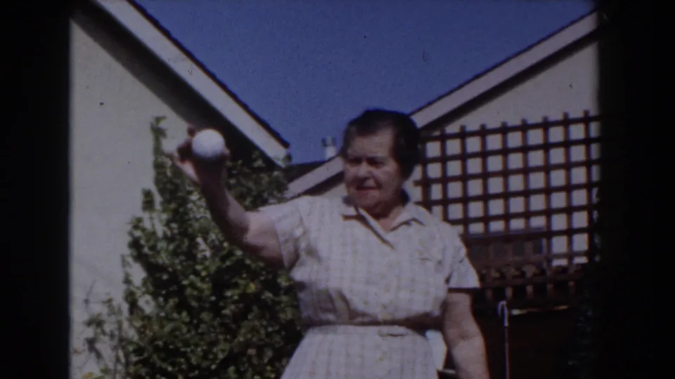 The older woman raises a white ball in the Glendale backyard, framed by a trellis, greenery, roofline, and blue sky.