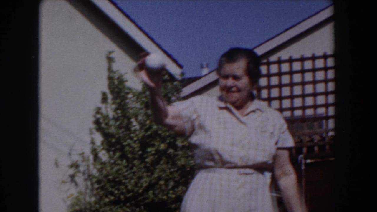 Older woman in a checked dress raises a white ball in a sunny backyard.