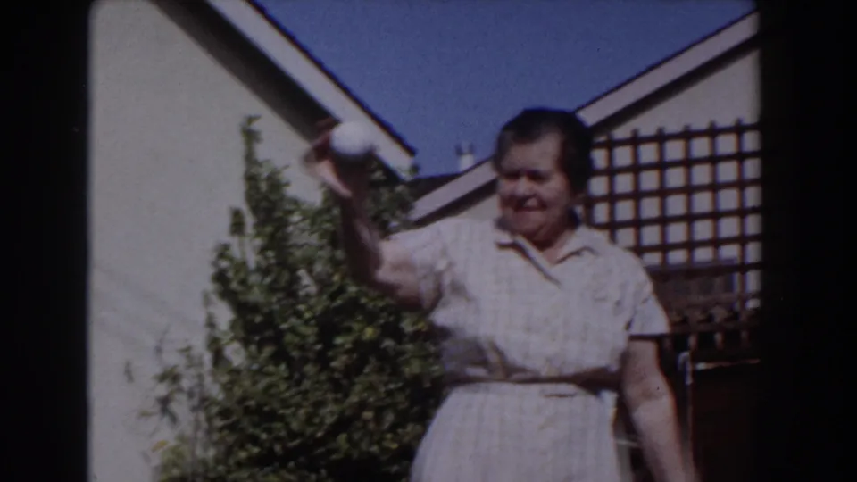 In Glendale, California home-movie footage from 1962, an older woman in a checked dress raises a white ball beside shrubs and a wall.