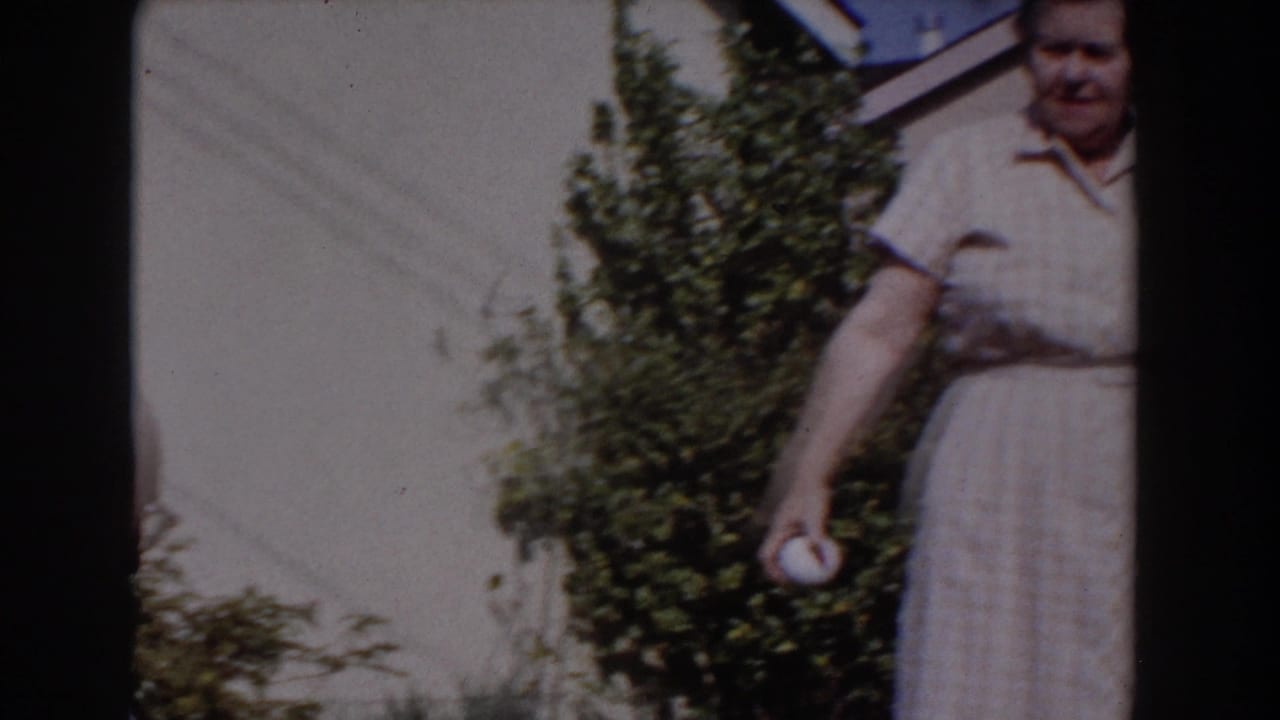 Older woman stands at the edge of frame holding a white ball near garden shrubs.