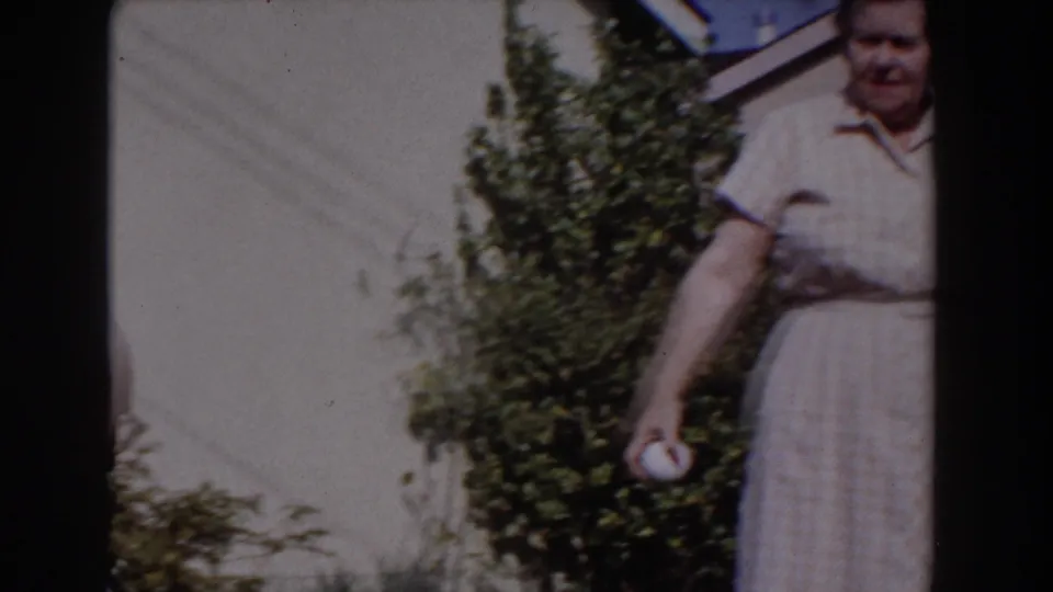 The older woman remains partly framed at the right, holding a white ball in the sunny Glendale backyard scene.