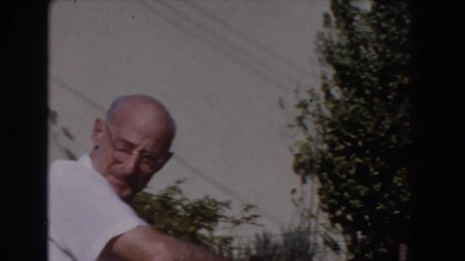 An older man in glasses and a white shirt leans forward near shrubs in the 1962 Glendale backyard home-movie scene.