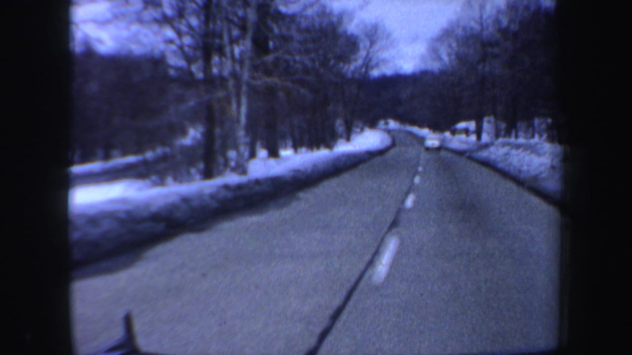 Snow-lined road seen through a car windshield with bare trees and a distant car ahead.