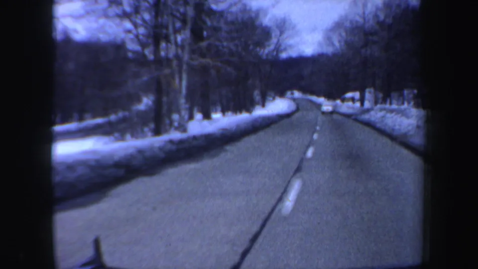 1962 New Hampshire 8mm frame of a winter road from inside a moving car, with snowbanks, bare trees, and a car ahead.