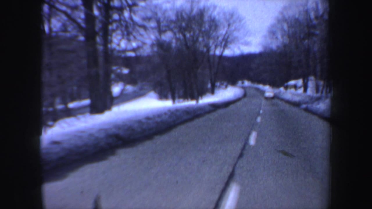 Curving winter road with snowbanks, bare trees, and a white car in the distance.