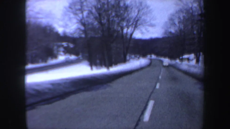 Soft 8mm view of a New Hampshire winter drive, showing a snow-edged road, bare trees, and low hills ahead.