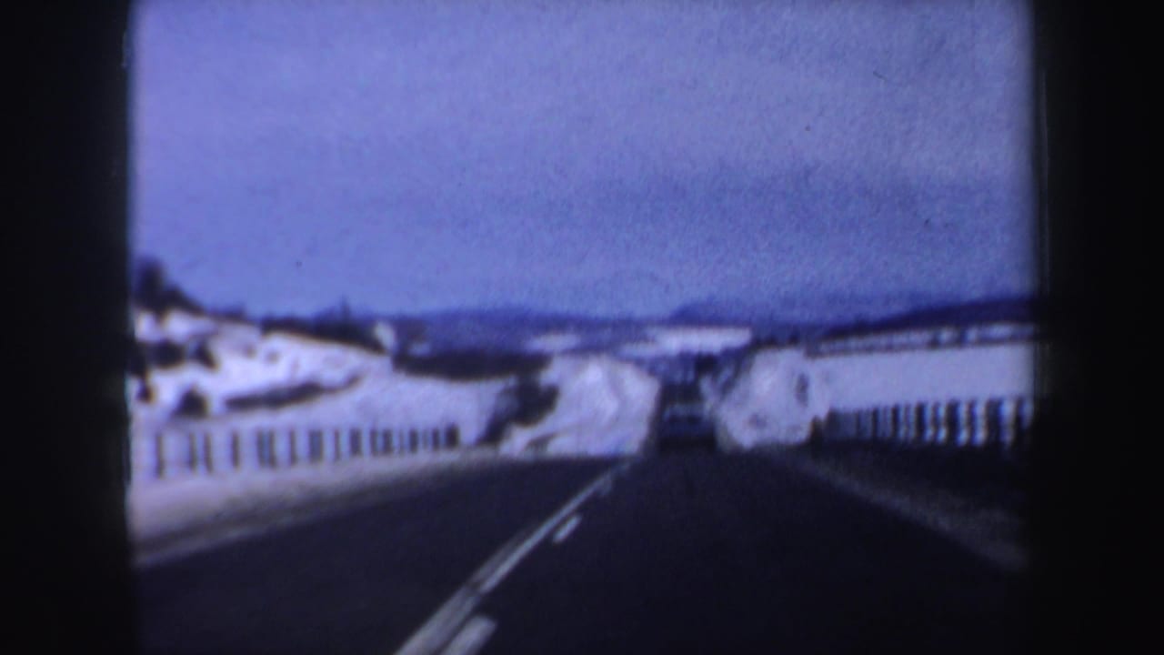 Snow-lined highway seen from a moving vehicle with guardrails and a car ahead.