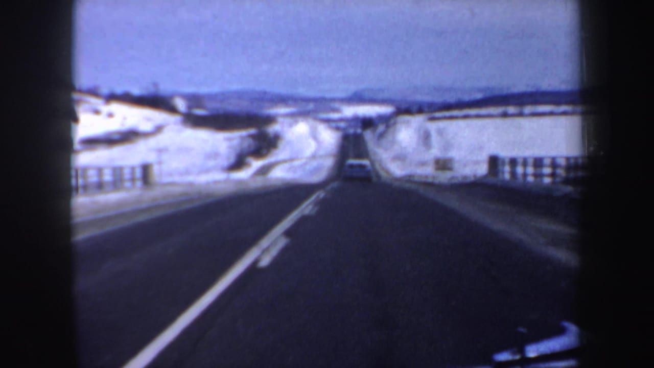 Highway stretches between snowbanks with guardrails and a vehicle in the distance.