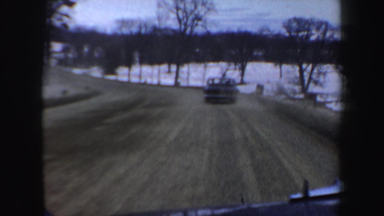 1962 New Hampshire 8mm frame from a moving vehicle approaching a frozen lake area, with a car ahead, bare trees, snow, and dashboard edge visible.