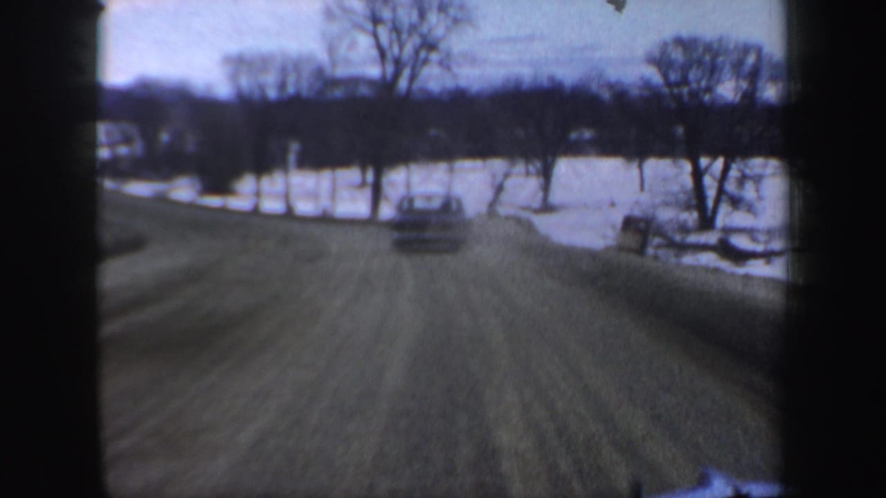 Rural road curving beside snow and a frozen lake, with a car ahead and bare trees in the distance.
