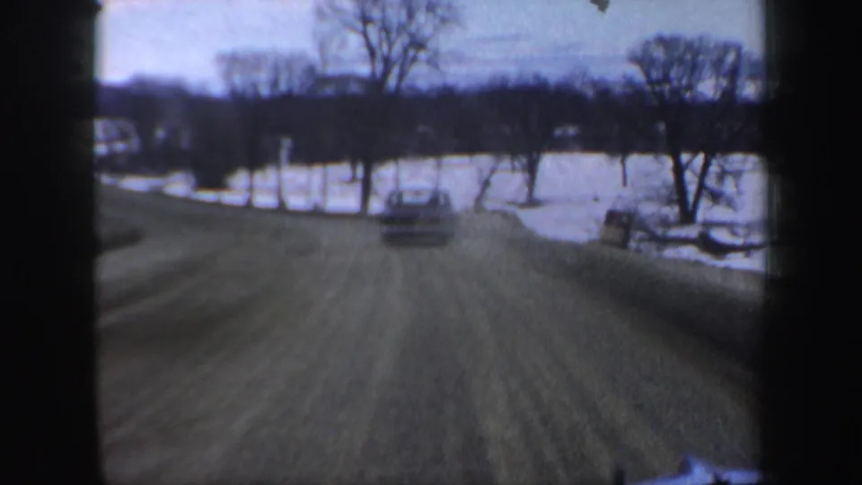 1962 New Hampshire road view beside a frozen lake or snowy shoreline, with a car ahead, winter trees, and blurred windshield framing.