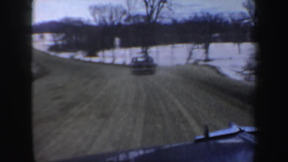 Home-movie frame from a moving vehicle on a New Hampshire winter road, showing snow, bare trees, a car ahead, and a frozen lakeside area.